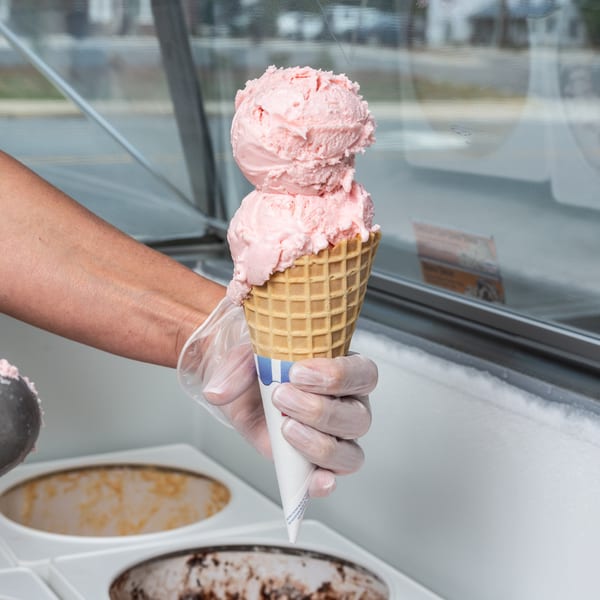 Gloved hand holding a waffle cone with two scoops of pink strawberry ice cream over an ice cream freezer.