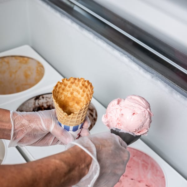 Gloved hands scooping pink ice cream into a waffle cone from a freezer.