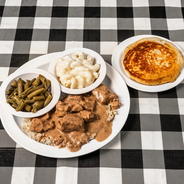 A plate of beef tips and rice, green beans, macaroni and cheese, and a pancake on a checkered tablecloth.
