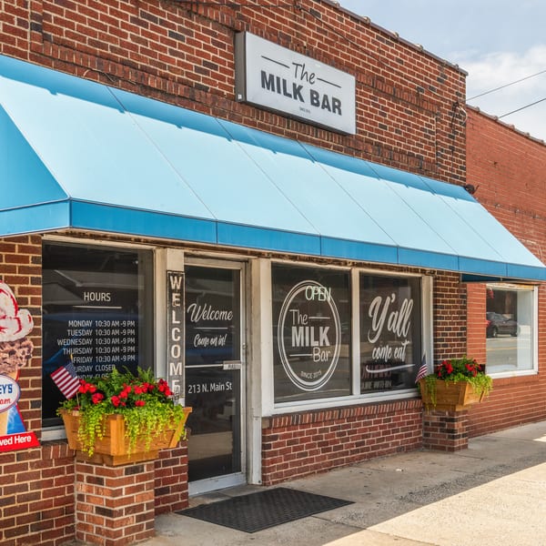 The Milk Bar storefront with a blue awning, brick facade, and window signs.