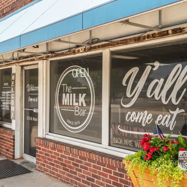 Exterior of The Milk Bar, a brick building with a blue awning and "Y'all come eat" on the window.
